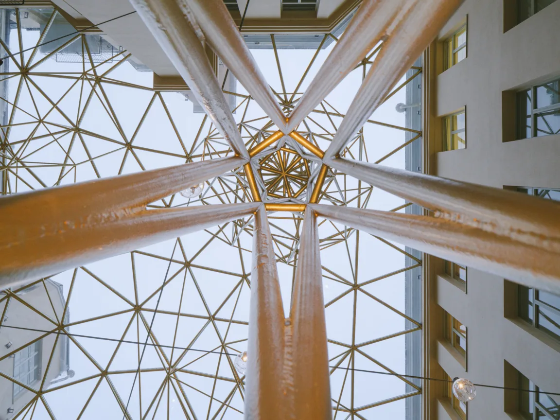 View of a geometric architectural structure from below, showcasing a network of golden beams and glass panels, with a clear sky visible through the design. The image highlights modern building aesthetics and intricate construction details.