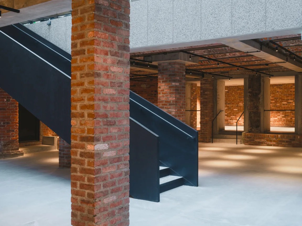 Modern industrial interior featuring a black staircase and exposed brick columns. The spacious area is illuminated with soft lighting, highlighting the blend of concrete and brick materials. Ideal for showcasing architectural design and urban aesthetics.