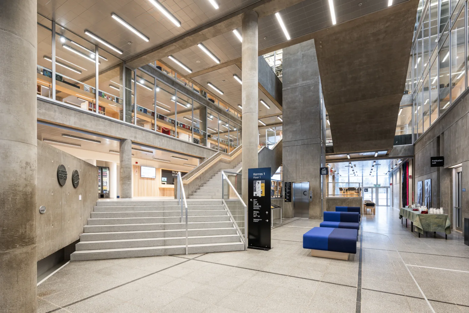 Modern library interior featuring spacious staircases, concrete pillars, and large windows allowing natural light. The design includes seating areas and informational signage, promoting a welcoming and functional study environment.
