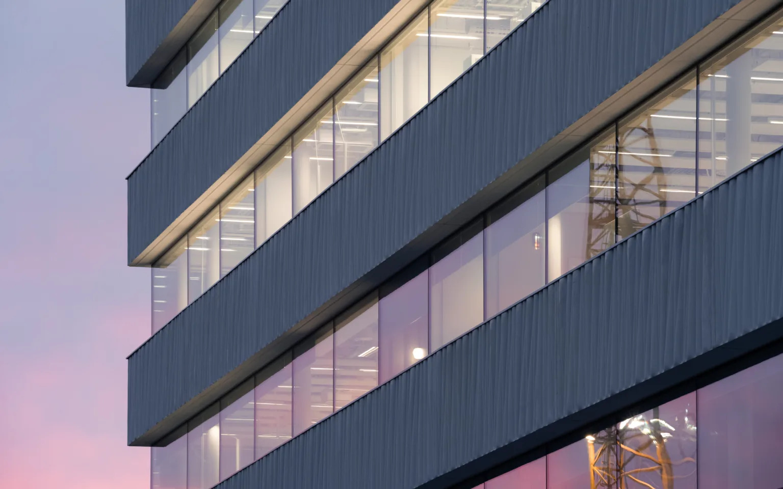 Modern office building facade with large glass windows and layered architectural design, illuminated against a colorful sunset sky.